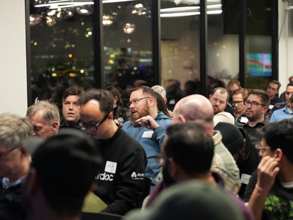 A candid photograph captures a large, seated audience at an indoor evening event, where one bearded man in the foreground is speaking into a microphone or asking a question while others listen. Large windows behind the audience reveal the illuminated cityscape at night.