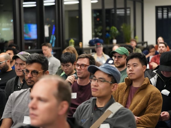 A candid photograph captures a diverse audience seated closely together during an evening indoor event, with one man in the center foreground holding a microphone, likely asking a question.