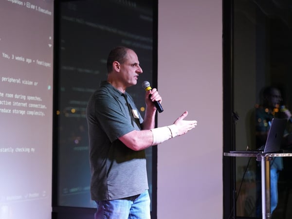 A man in a green polo shirt and jeans is giving a presentation, speaking into a microphone while gesturing toward a large projection screen displaying technical bullet points.