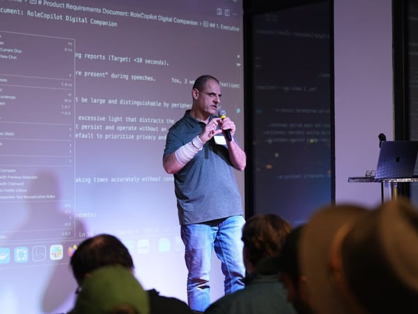 A man is giving a technical presentation on stage, holding a microphone, with a large projection screen behind him displaying a product requirements document titled 'RoleCopilot Digital Companion.' Several audience members are visible in the dark foreground.