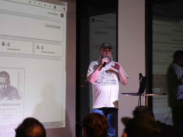 A photograph taken indoors shows a bearded man wearing a cap and a white t-shirt giving a presentation, holding a microphone and gesturing while standing in front of a projected screen displaying a user interface.