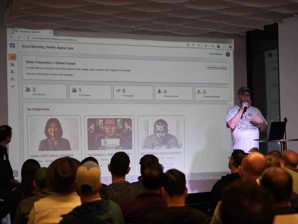 A man wearing a cap and glasses is giving a presentation using a microphone in front of a large projection screen displaying a web application dashboard to a seated audience in a dimly lit indoor space.