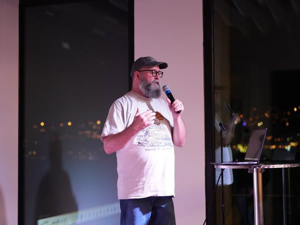 A photograph captures a bearded man wearing a baseball cap and glasses, speaking into a microphone while giving a presentation indoors at night. Behind him is a large window offering a view of a city skyline illuminated by bokeh lights.