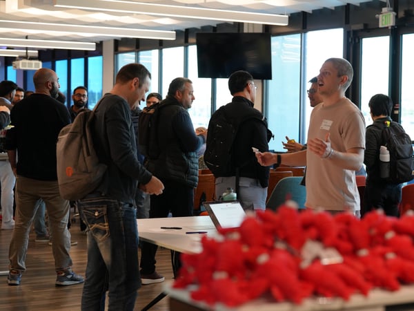 A group of people are seen socializing and networking at an indoor event, with a blurred pile of red stuffed toys in the foreground.