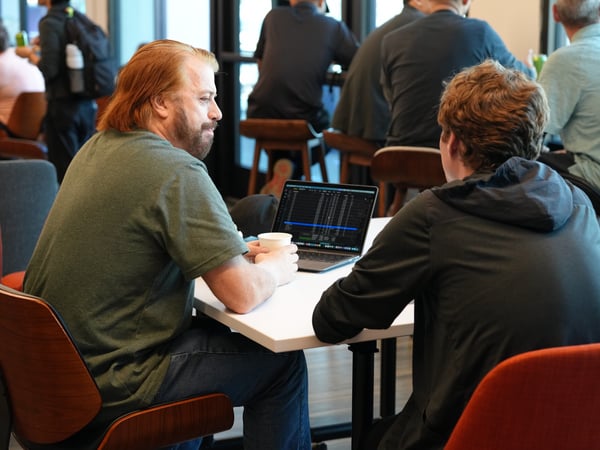 Two men are seated at a white table in a coworking space or cafe, engaged in a discussion with an open laptop between them.