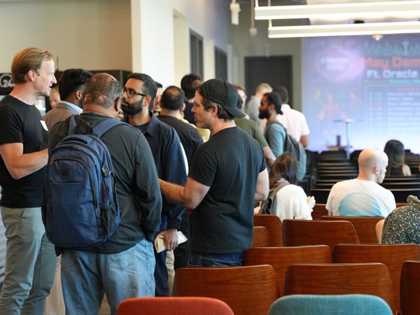 A group of people are standing and socializing in an indoor event space, with rows of empty wooden chairs and a presentation screen visible in the background.