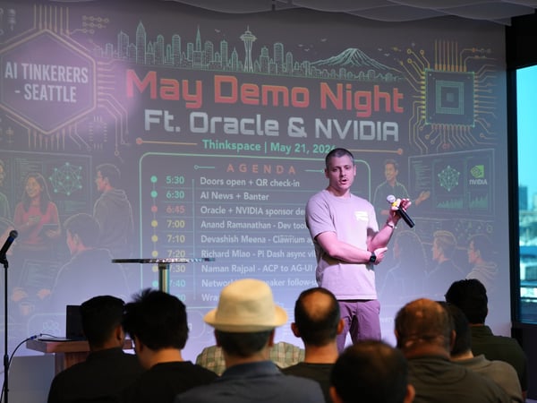 A photograph showing a speaker presenting on stage at an 'AI Tinkerers - Seattle' event, with an audience in the foreground and a large projection screen displaying the event's agenda behind him.