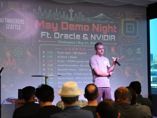 A photograph capturing a presenter speaking into a microphone on stage during a tech event, with an audience in the foreground and a large projector screen displaying the 'May Demo Night' agenda behind him.
