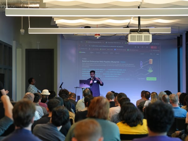 A presenter stands on stage speaking into a microphone in front of a projection screen displaying an NVIDIA website about building an enterprise RAG pipeline, while an audience watches from their seats.