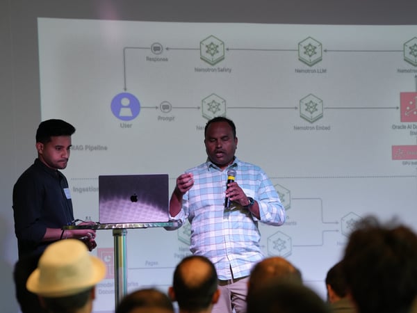 A photograph showing two men presenting on a stage in front of a projection screen displaying a technical system architecture diagram. One man is speaking into a microphone while the other stands next to a laptop on a high table.