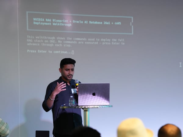 A man stands at a high table presenting a slideshow on a projection screen behind him, speaking into a microphone with a laptop in front of him.