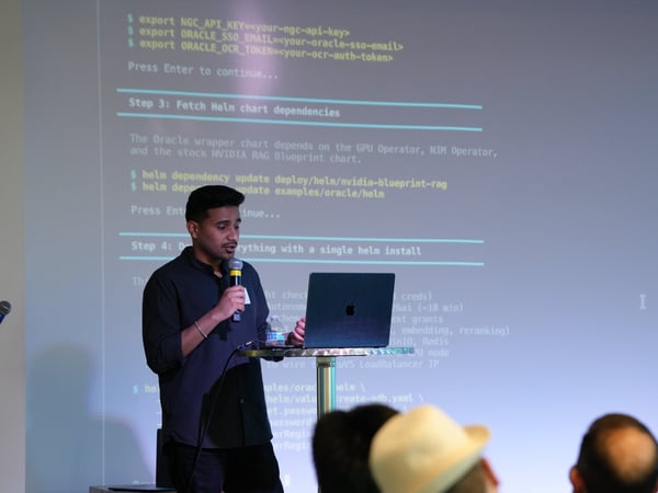 A presenter stands at a podium with a laptop and microphone, delivering a technical presentation with command-line instructions projected on a screen behind him.