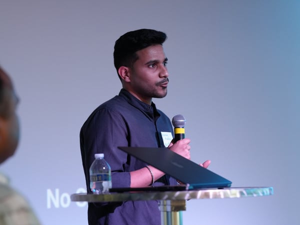 A young man speaks into a microphone during a presentation, standing behind a high table with a laptop and a water bottle.