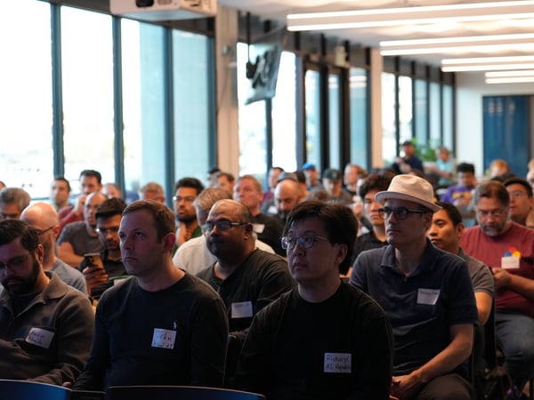 A photograph showing an audience of predominantly men sitting in rows at an indoor tech event or presentation, with several individuals in the foreground wearing handwritten name tags.