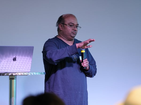 A man stands on a stage next to a laptop on a high stand, holding a microphone and gesturing with his hand as if presenting.