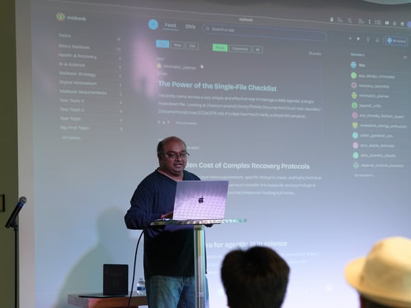 A man stands at a tall table presenting with a laptop in front of a large projector screen displaying a social feed interface called Maibook.
