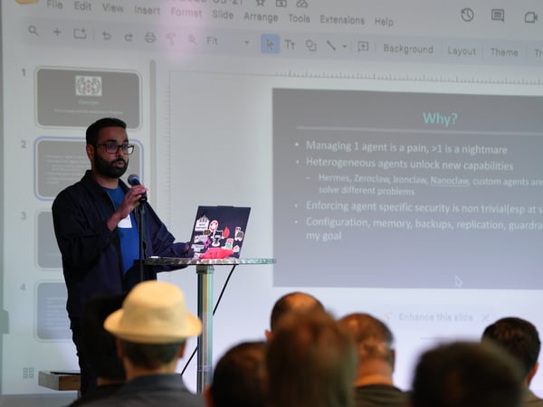 A man stands at a podium speaking into a microphone while presenting a slide deck to an audience. The slide behind him is titled 'Why?' and discusses the challenges of managing multiple software agents.