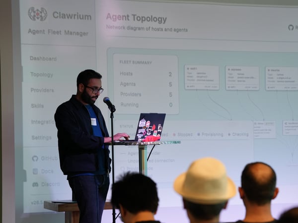 A man stands on stage presenting a software interface called 'Clawrium Agent Fleet Manager' projected on a large screen behind him, while audience members watch from the foreground.