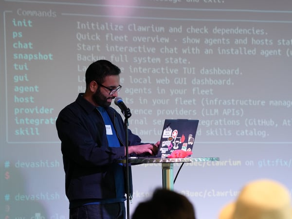 A man stands on stage at a high table, typing on a sticker-covered laptop during a presentation. Behind him, a large screen displays terminal commands and instructions for a tool named Clawrium.
