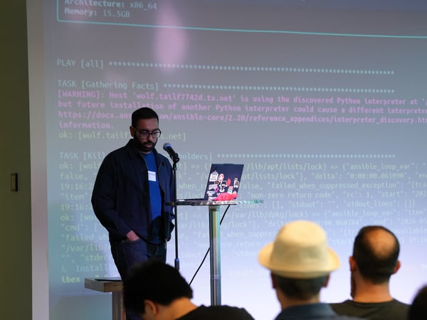 A man stands at a podium presenting a technical talk with a microphone, in front of a large projection screen displaying terminal output and code logs, while an audience watches from the foreground.