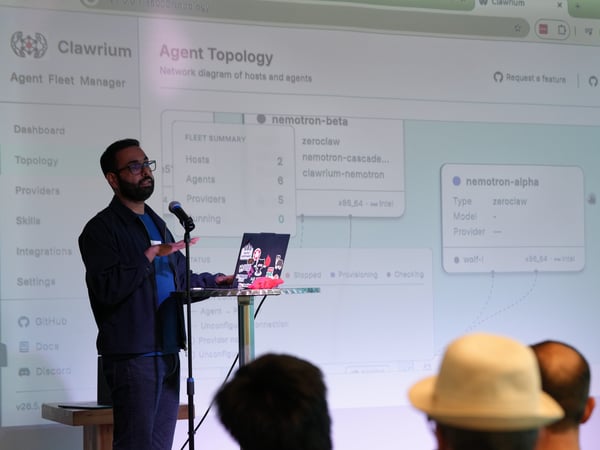 A man stands on stage presenting a network topology diagram projected on a large screen behind him, speaking into a microphone with his laptop open on a stand.