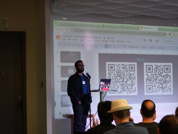 A man stands at a microphone giving a presentation at a tech event, with a projector screen behind him displaying a Google Slides presentation with two QR codes.