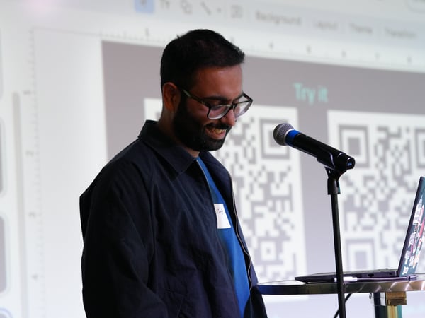 A man with glasses and a beard is smiling while standing at a podium with a microphone and laptop, presenting in front of a projection screen showing a slide with QR codes.