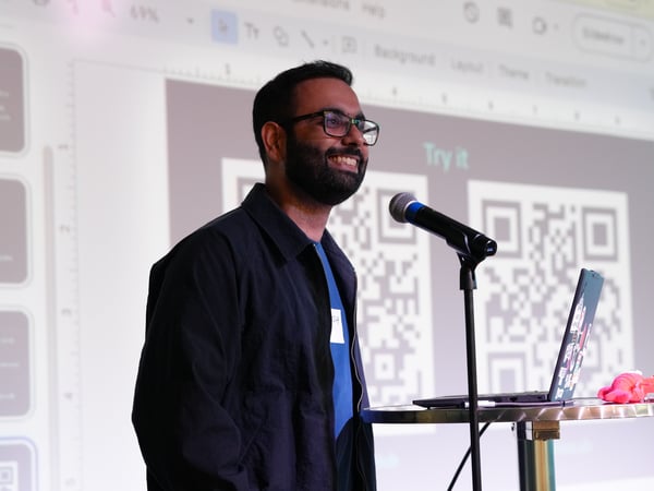 A man with glasses and a beard is smiling while standing at a high table with a laptop and a microphone, presenting in front of a projection screen showing QR codes.