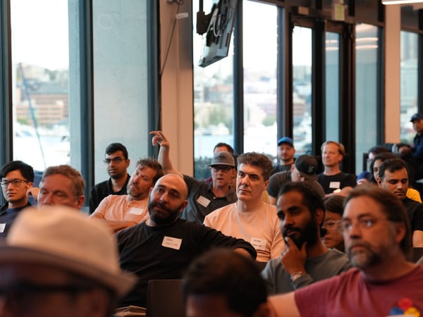 A photograph showing an audience of people sitting indoors and attentively listening to a presentation, with large windows in the background overlooking a waterfront city view.