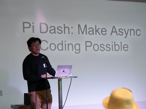 A man stands at a high table presenting with a laptop in front of a large projection screen displaying the text 'Pi Dash: Make Async Coding Possible'.