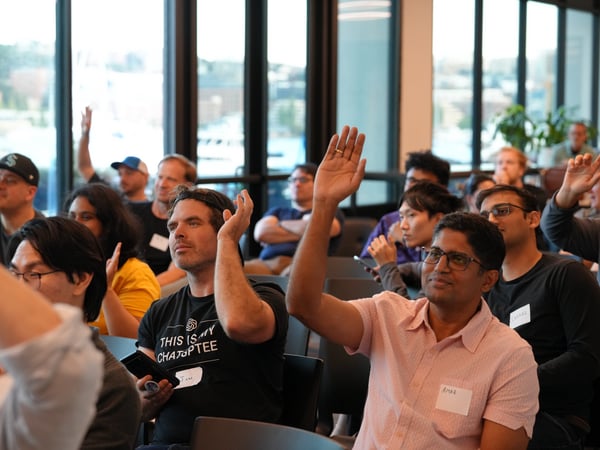 An indoor shot of an audience at a tech meetup or presentation, with several attendees raising their hands to ask questions or participate.