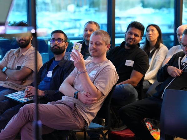 A group of people are sitting in chairs, attentively watching a presentation or event indoors. One man in the foreground is holding up a smartphone with a sticker on the back.