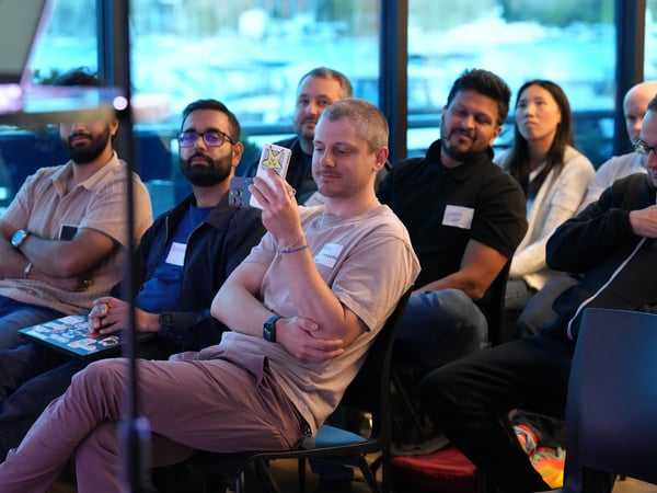 A photograph showing an audience of several people sitting in chairs indoors, listening to a presentation. One man in the foreground is holding up his smartphone, which has a star sticker on the back.