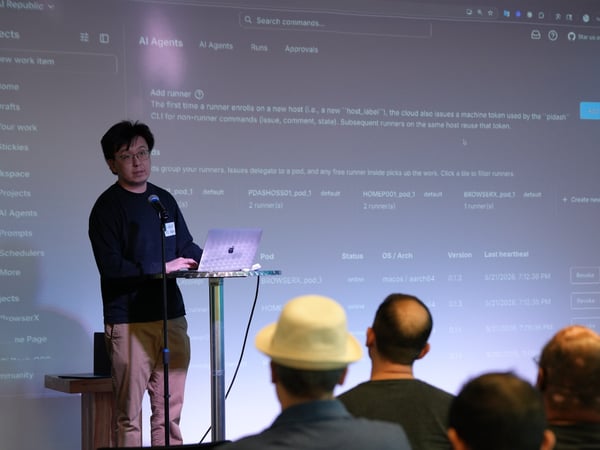 A man stands on stage giving a presentation, speaking into a microphone next to a high table with a laptop, in front of a large projection screen displaying a software dashboard.