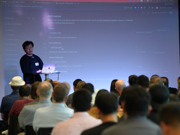 A man stands on a stage presenting a software interface on a large projection screen to a seated audience.
