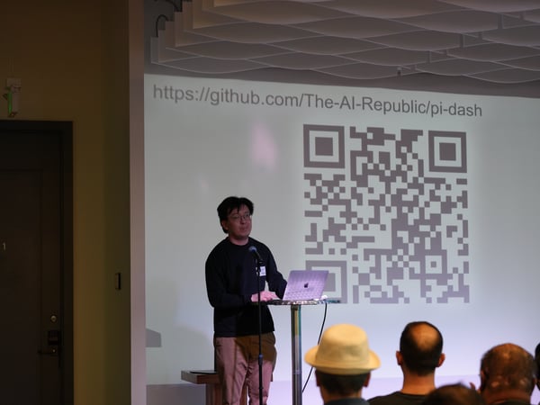 A man stands on a stage giving a presentation next to a laptop, with a large projector screen behind him displaying a QR code and a GitHub URL.