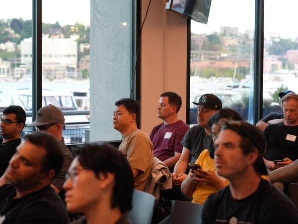 An indoor photograph showing an audience of people seated and listening to a presentation, with large windows in the background offering a scenic view of a waterfront and buildings.