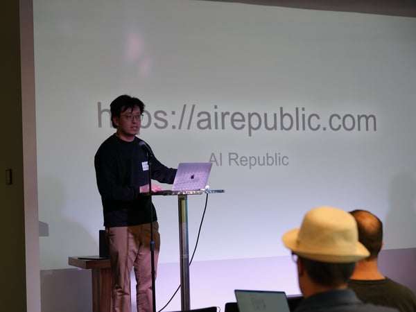 A man stands at a high table presenting with a laptop and microphone in front of a projector screen displaying a website URL.