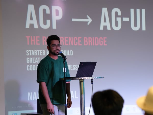 A young man wearing glasses and a green t-shirt is speaking into a microphone during a presentation, standing next to a laptop on a high stand with a slide projected behind him.
