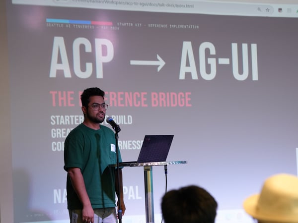 A photograph of a presenter standing at a podium with a laptop and microphone, delivering a talk in front of a projected slide titled 'ACP -> AG-UI THE REFERENCE BRIDGE'.