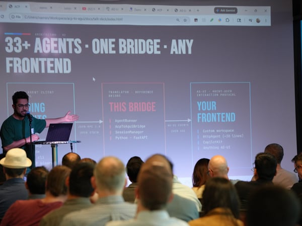 A man is giving a presentation on stage in front of a projection screen displaying a slide about an agent bridge architecture, with an audience watching from their seats.