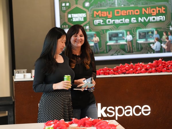 Two women stand and converse at a tech event, with a digital screen in the background advertising 'May Demo Night (Ft. Oracle & NVIDIA)'.