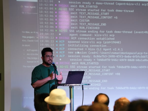 A man in a green shirt and glasses stands on stage presenting, speaking into a microphone next to a laptop. Behind him, a large screen displays scrolling terminal logs and code from a software execution.