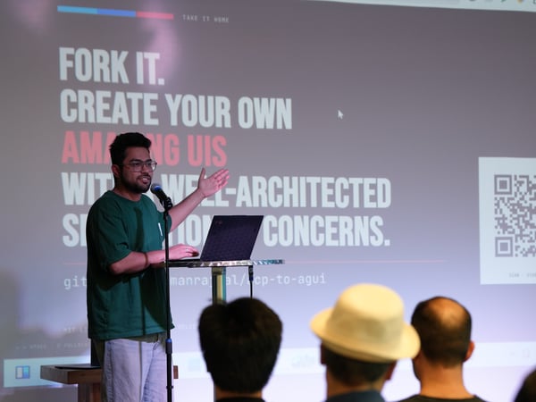 A man stands at a podium giving a presentation, gesturing towards a large projector screen displaying text and a QR code in front of an audience.