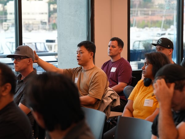 An indoor photograph showing an audience seated during a presentation or Q&A session, with a man in a tan t-shirt raising his hand to ask a question.