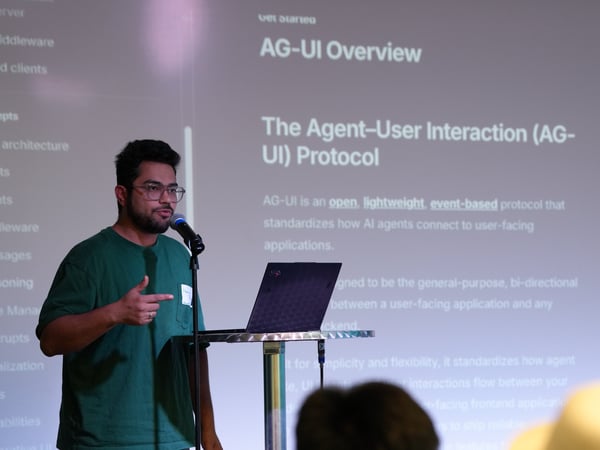 A man in a green t-shirt and glasses is giving a presentation at a podium with a laptop and microphone, in front of a screen displaying slides about the AG-UI protocol.