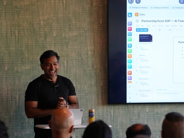 A man stands smiling next to a small table while presenting, with a large screen behind him showing a presentation slide.