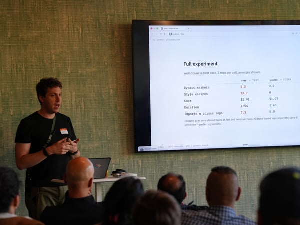 A presenter stands next to a large display monitor showing experimental data to a seated audience.