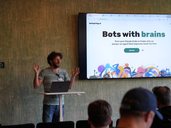 A presenter stands at a standing desk next to a large display showing a website titled 'Bots with brains'. Several audience members are visible in the foreground.
