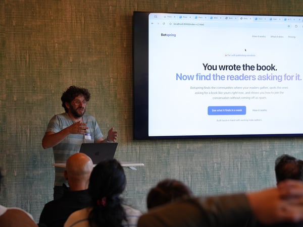 A presenter stands next to a laptop on a stand, gesturing toward an audience in front of a large monitor displaying a website mockup.